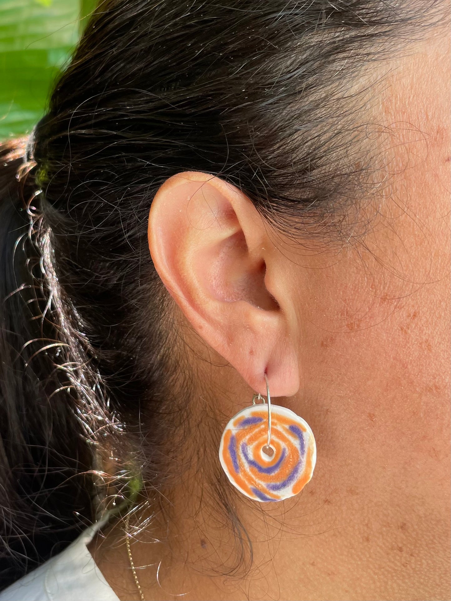 Closeup of woman wearing orange and lavender hoops earrings. Earrings are in a simple rose pattern with orange and lavender brush strokes making loose circular patterns. Woman has tan skin and dark brown hair pulled back in a ponytail. A vivid green bird of paradise leaf is in the background and the sun is shining on her hair.