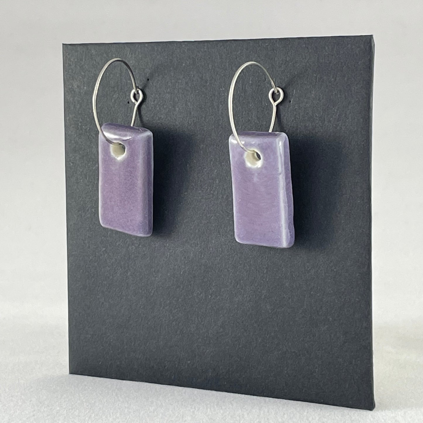 Side view of a pair of rectangular shaped porcelain hoop earrings in purple. On sterling silver earrings hooks.