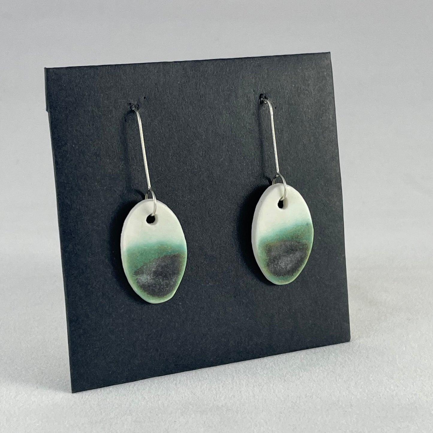 Pair of emerald green and white oval shaped porcelain earrings. Top half white and bottom half textured green. Sterling silver hooks. Side view