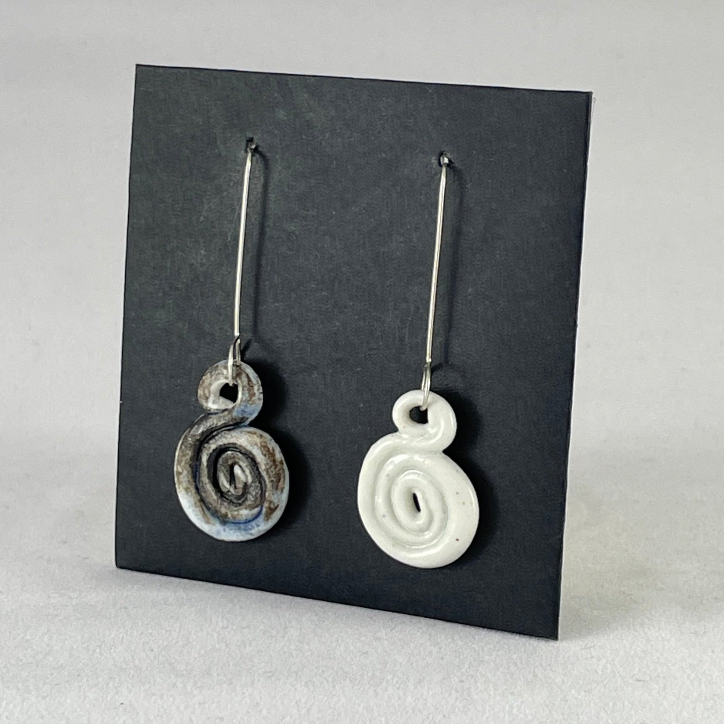 A pair of hand coiled porcelain earrings. One earring is white and the other is blue and black. Photographed on a black background. On sterling silver earrings hooks. Side view