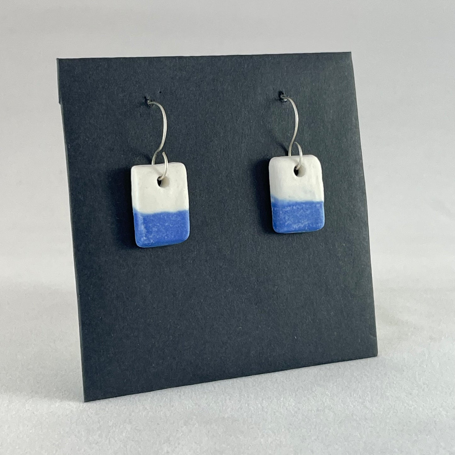 A pair of rectangular porcelain earrings in half white and half ultramarine blue. Photographed on a black background. Side view
