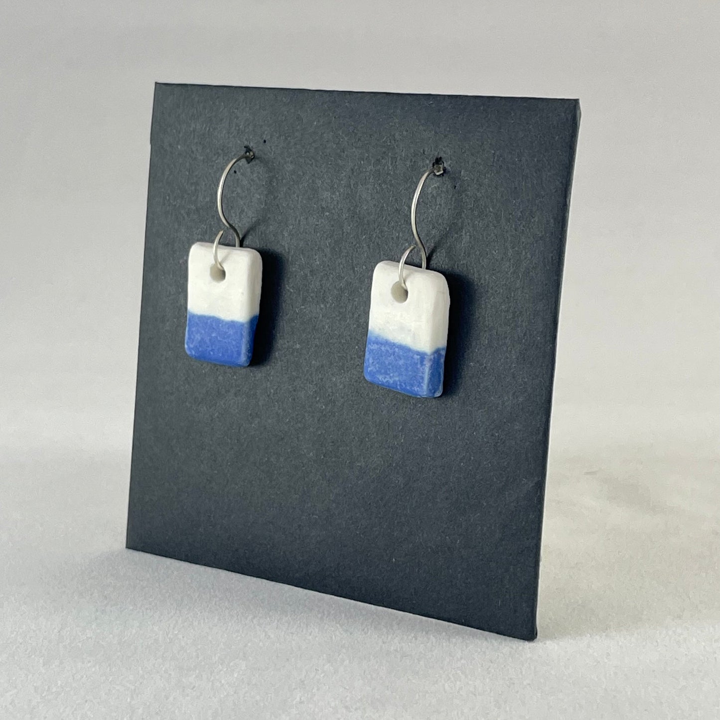 A pair of rectangular porcelain earrings in half white and half ultramarine blue. Photographed on a black background. Side view