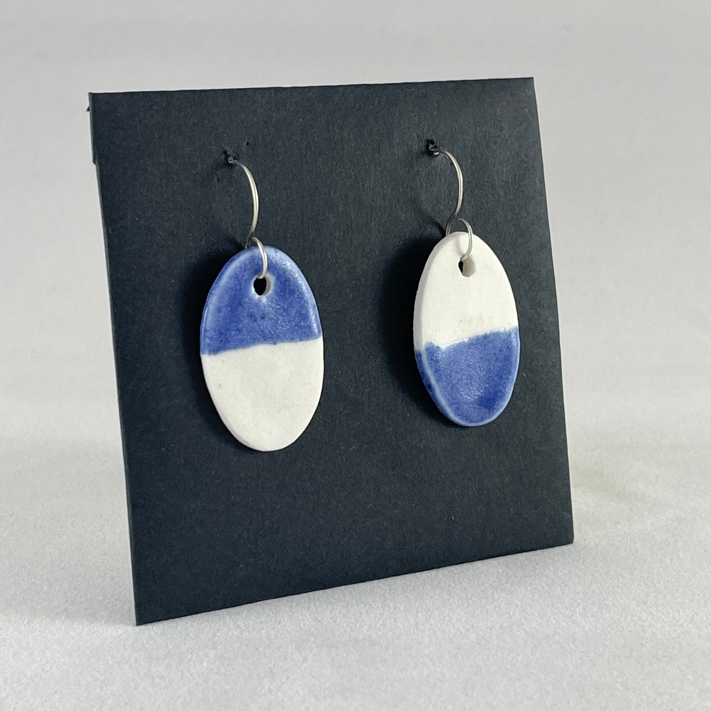 A pair of oval shaped porcelain earrings in half white and half ultramarine blue. Photographed on a black background. Side view