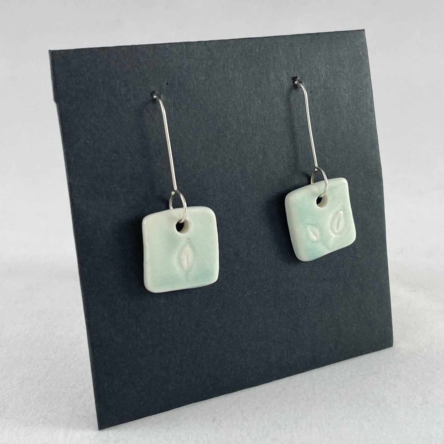 A pair of mint green square porcelain earrings on a black background. On sterling silver earrings hooks. Side view facing right