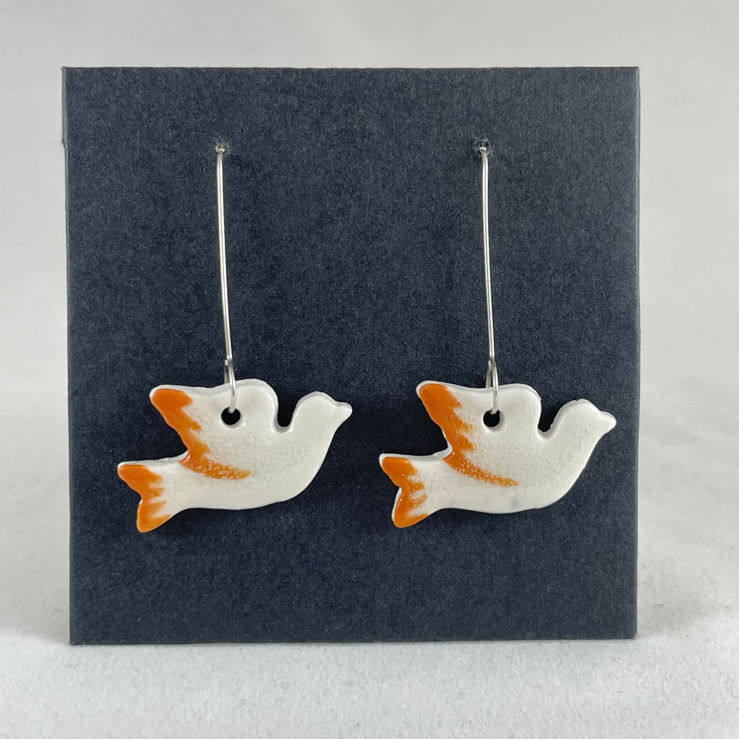 A pair of white and orange porcelain earrings in the shape of flying dove on black card.