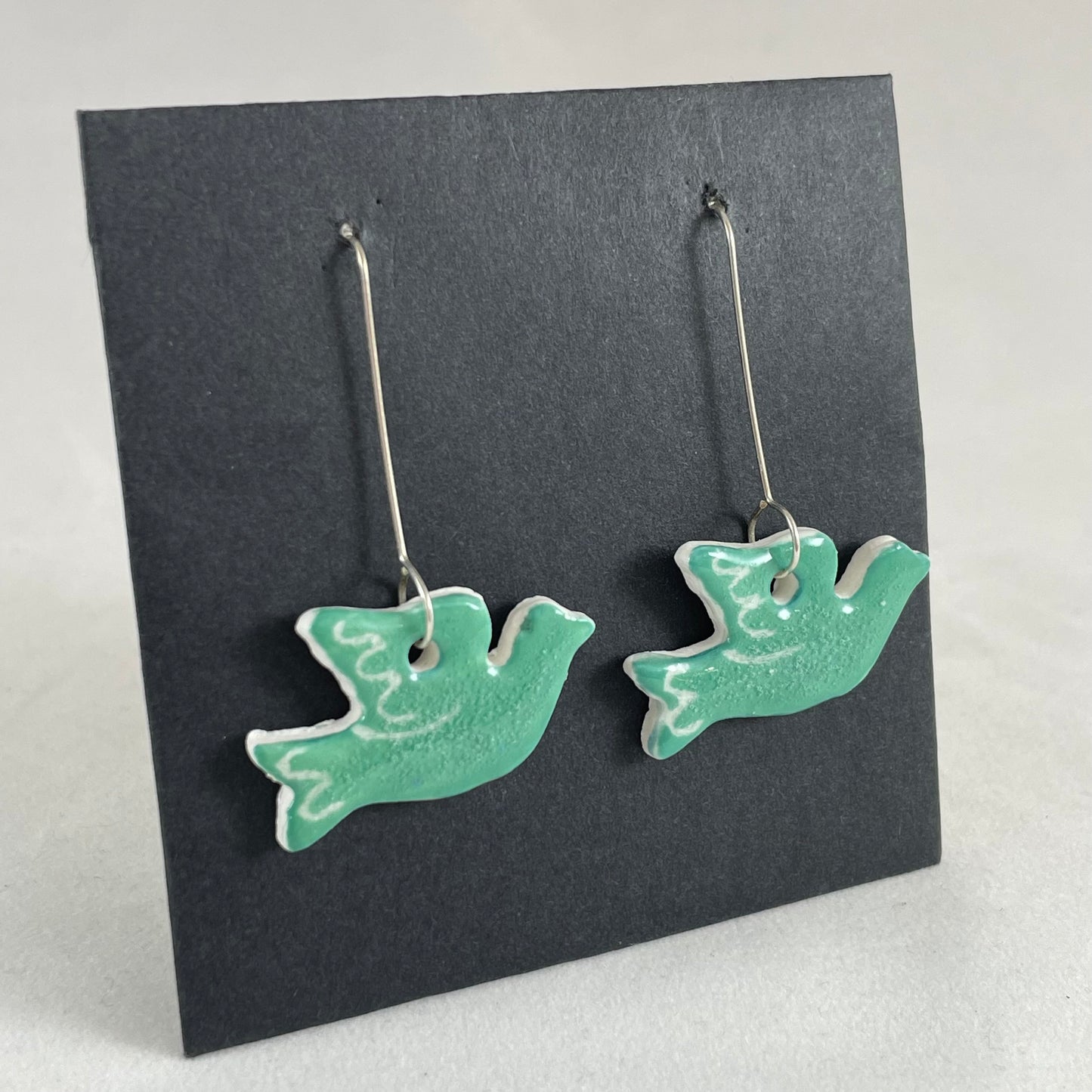 A pair of green porcelain earrings in the shape of flying dove on a black background. Side view