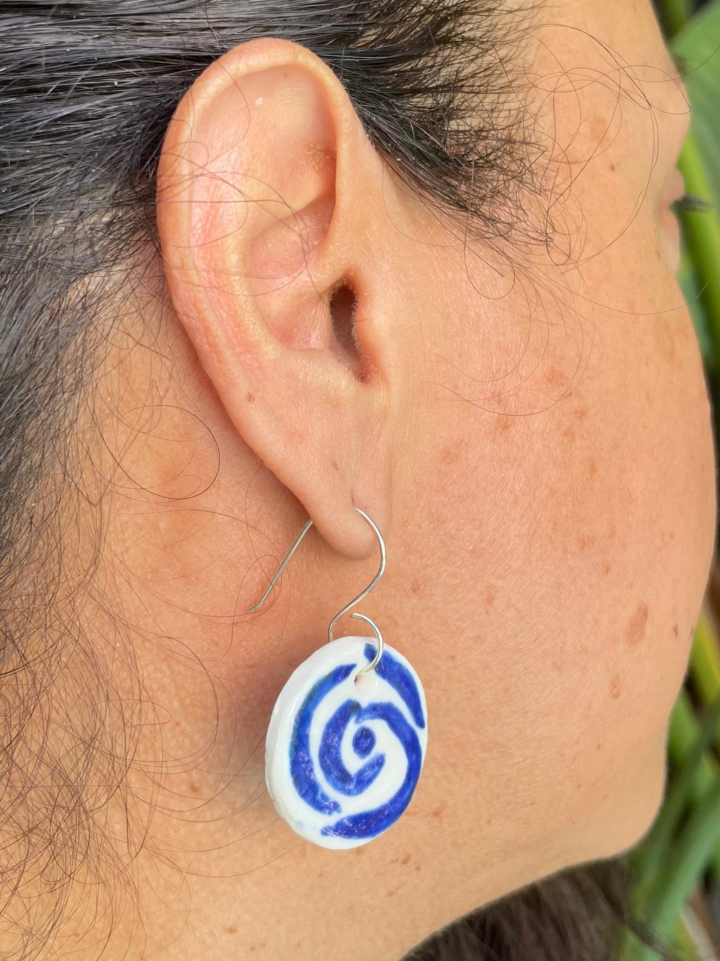 Closeup of woman with tan skin wearing white and blue circular earrings with rose pattern. Woman has her dark brown hair is pulled back in ponytail.