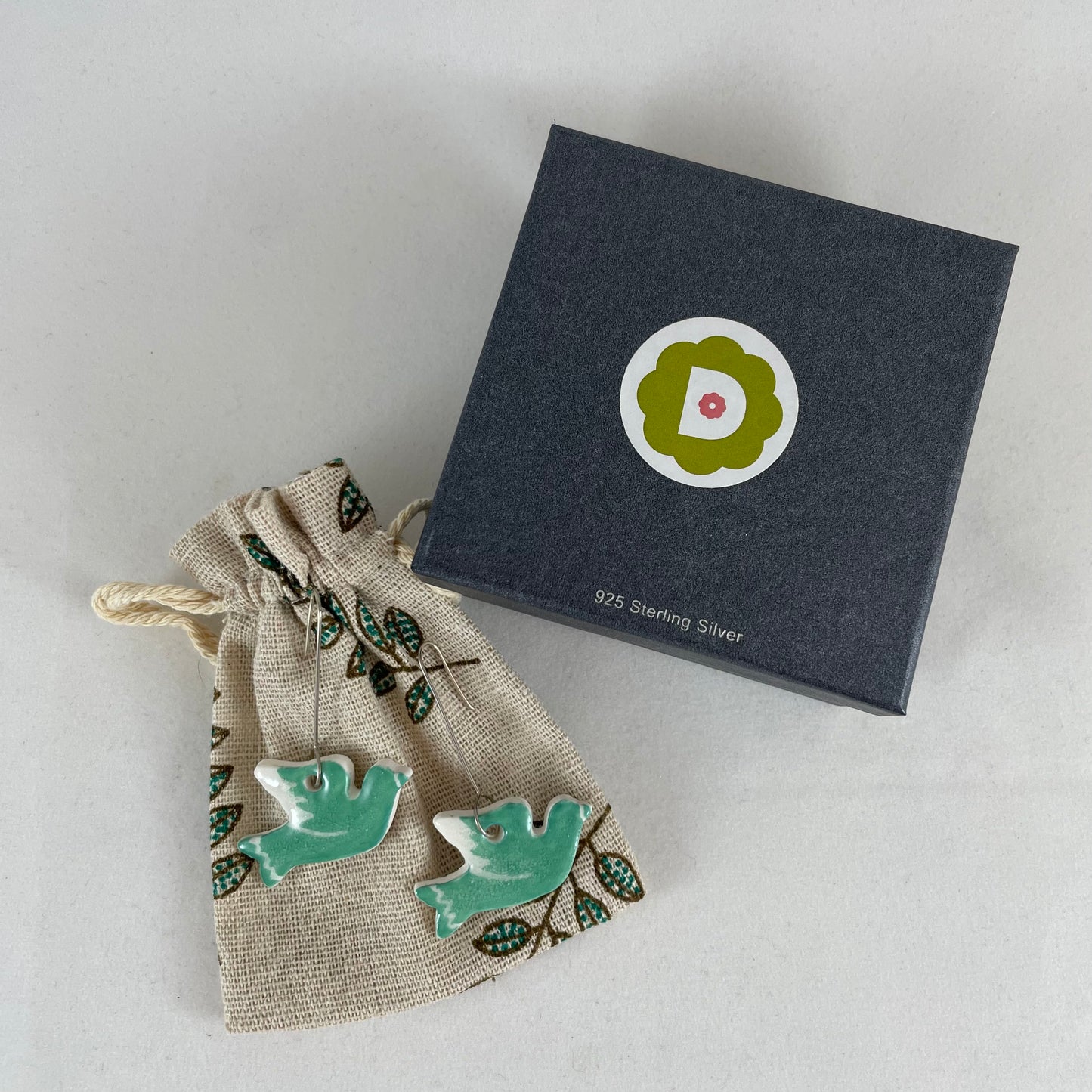 A pair of green porcelain earrings in the shape of flying dove. Photographed on a linen pouch next to jewellery box.