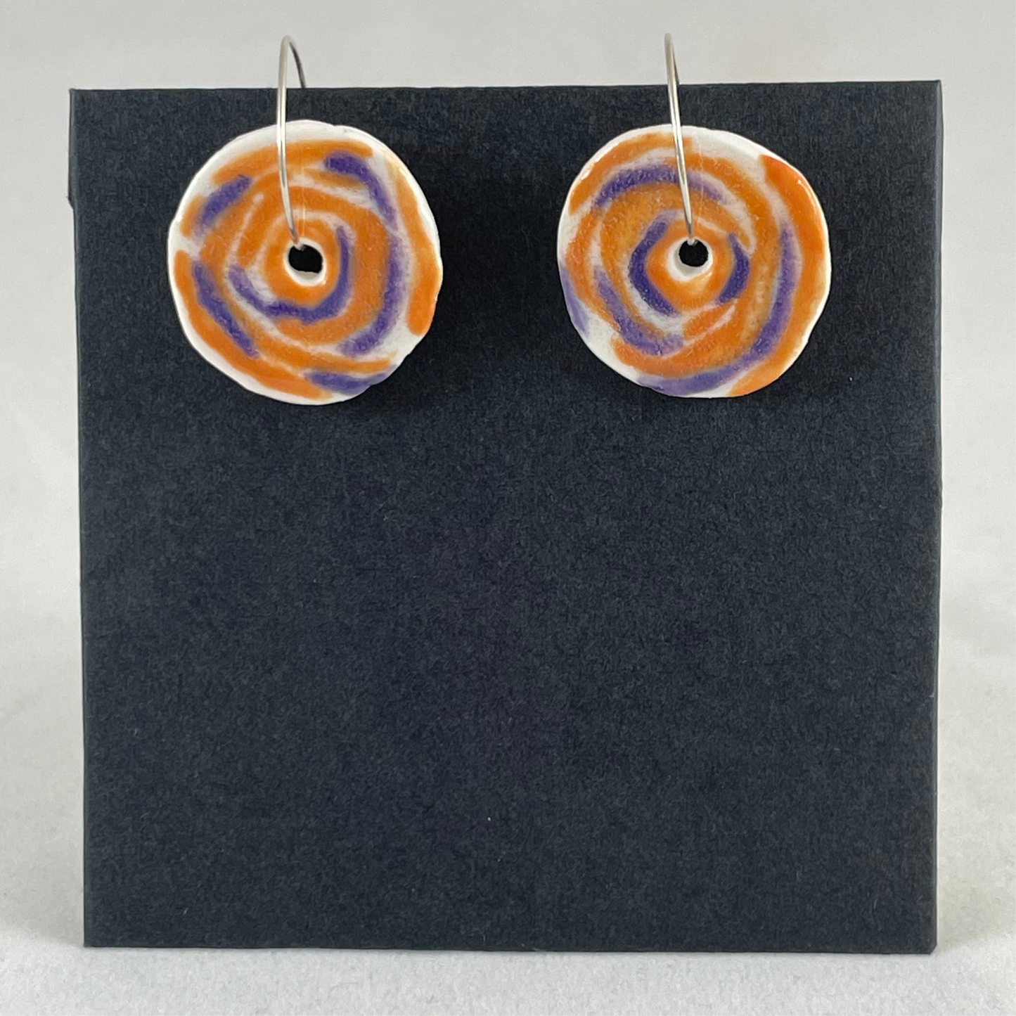 A pair of lavender and orange porcelain earrings on black card. Simple rose pattern with loosely painted orange and lavender circles and small areas of white porcelain.