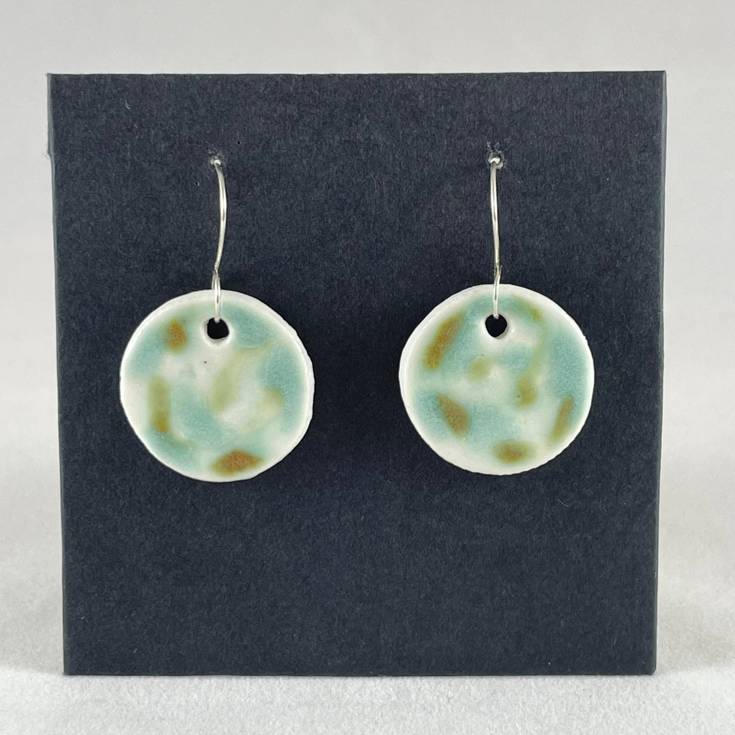 A pair of mint, olive green and white porcelain earrings hanging on black card. Splodges of glossy glaze look vaguely like a blurry camouflage pattern.