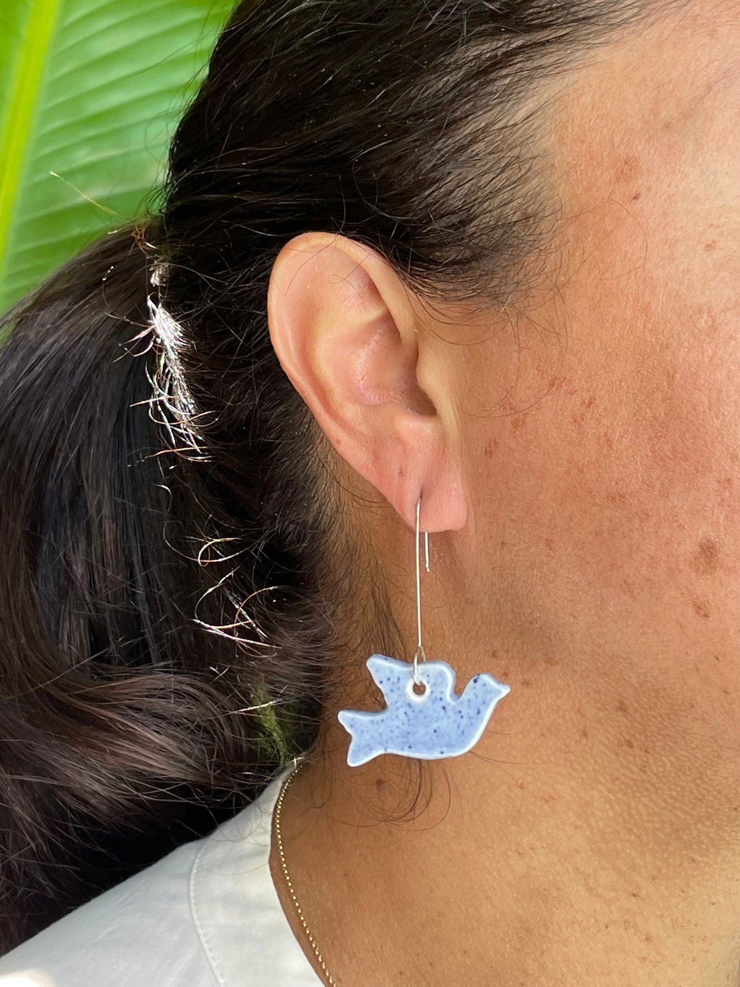 Closeup of woman wearing pale blue dove earrings. Woman has olive skin and her dark brown hair is pulled back in ponytail. Sun is shining in garden
