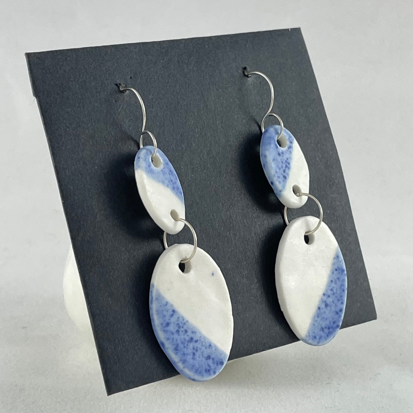 A pair of handmade porcelain earrings on a black background. Asymmetrical glaze pattern in blue and white on oval pendants. Side view