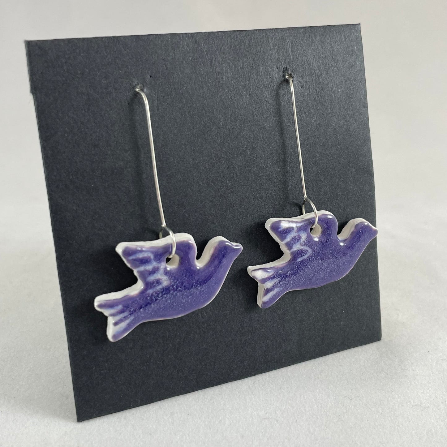 A pair of lavender porcelain earrings in the shape of flying dove on a black background. Side view