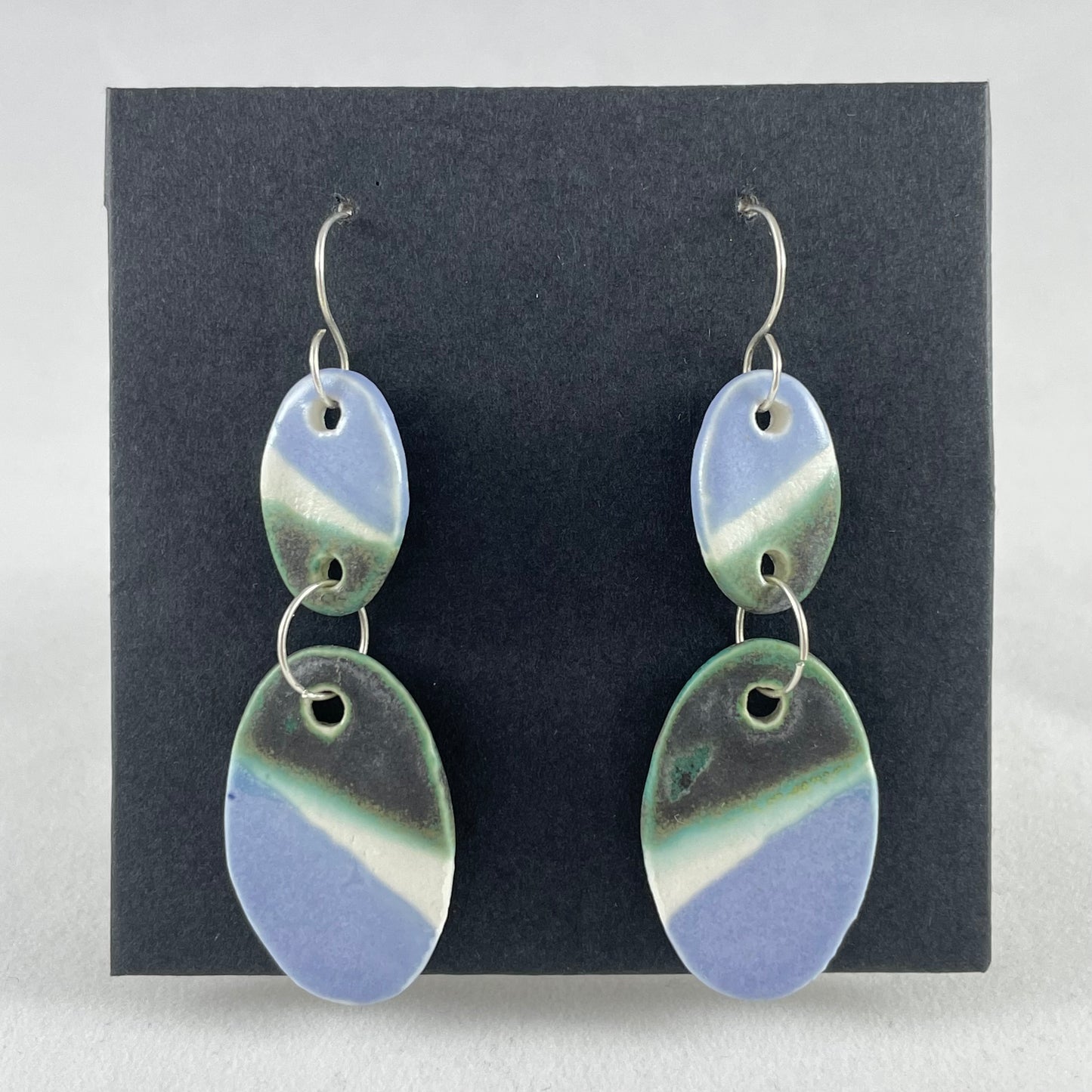 A pair of handmade porcelain earrings on a black background. Asymmetrical glaze pattern in very dark green, light blue and white on oval pendants.