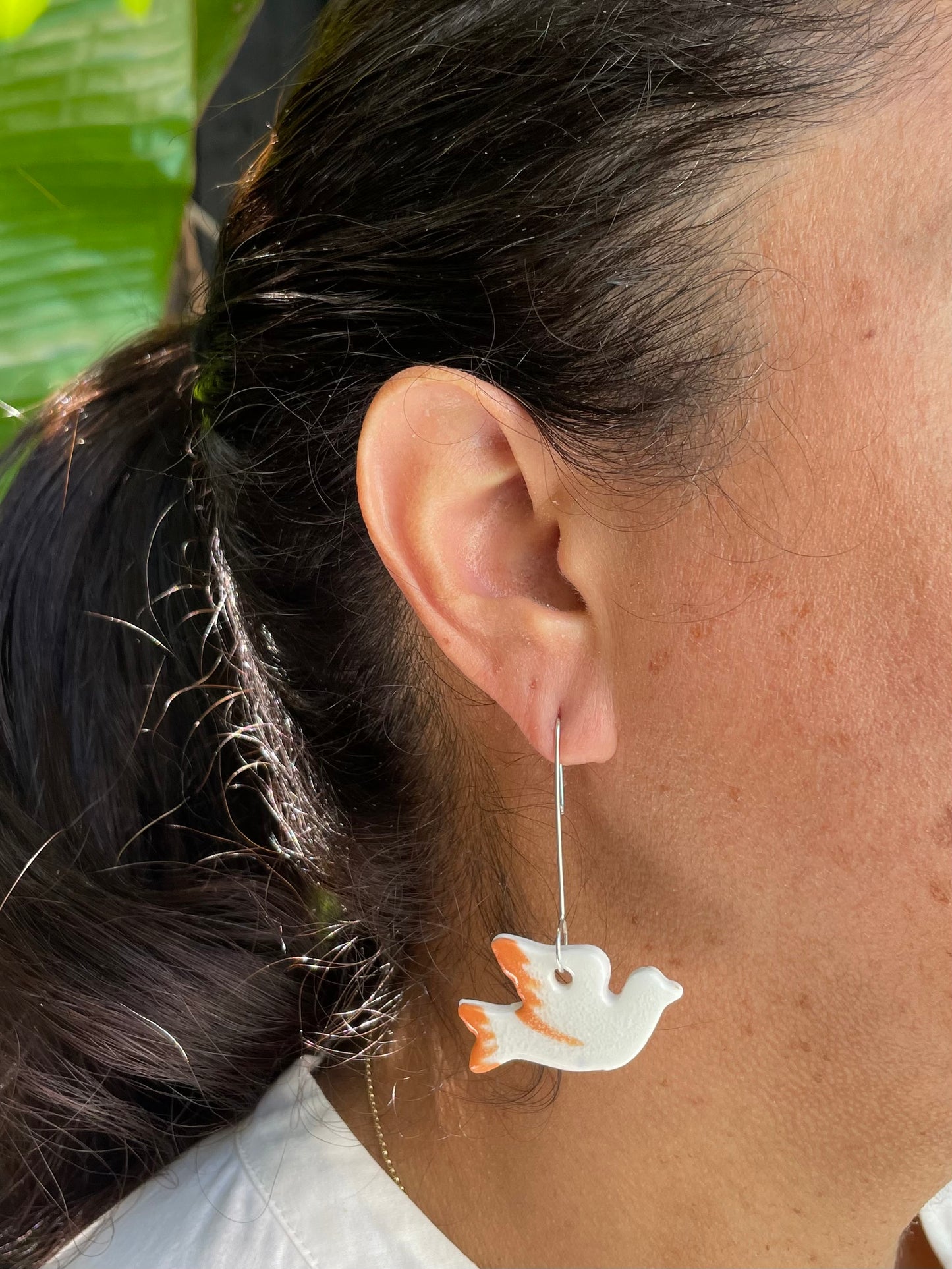 Closeup of woman with tan skin wearing white and orange dove earrings. Woman has her dark brown hair is pulled back in ponytail. A bright green bird of paradise plant is partially visible in the background