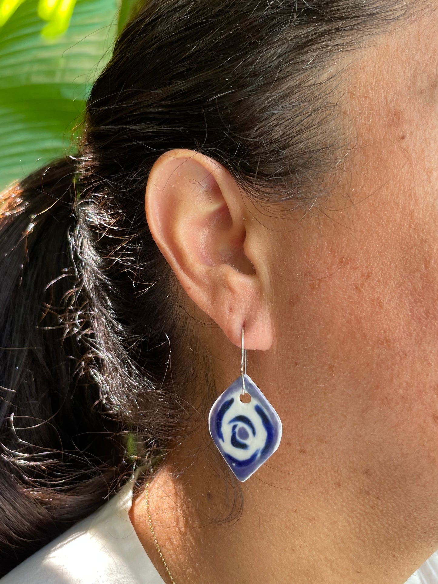 Closeup of woman with tan skin wearing lavender, blue and white pointed oval shape earrings with rose pattern. Woman has her dark brown hair is pulled back in ponytail. A bright green bird of paradise plant is partially visible in the background