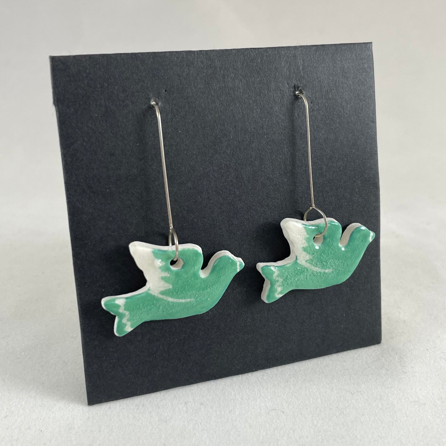 A pair of green porcelain earrings in the shape of flying dove on a black background. Side view