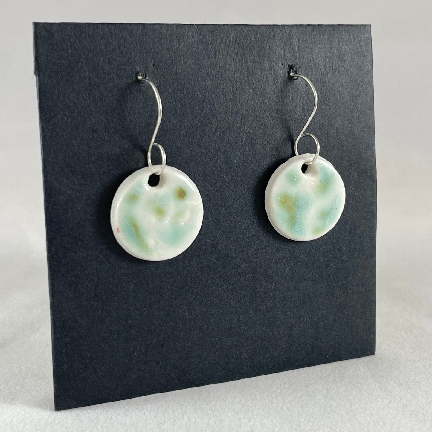 A pair of mint, olive green and white porcelain earrings hanging on black card. Photographed from left hand side showing slim earring profile.