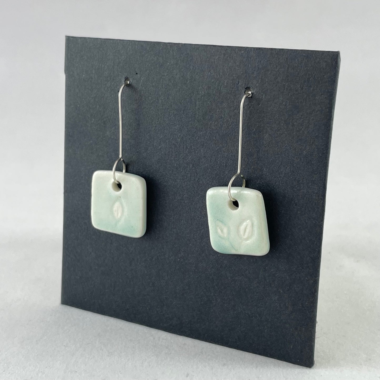 A pair of mint green square porcelain earrings on a black background. On sterling silver earrings hooks. Side view facing left