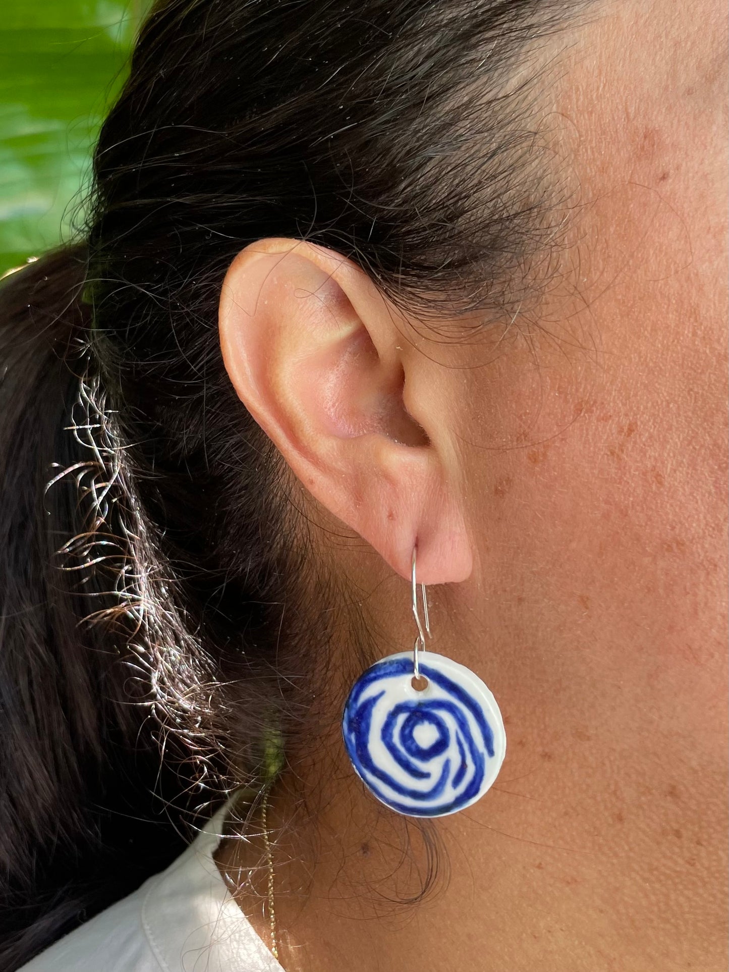 Closeup of woman with tan skin wearing white and blue circular earrings with rose pattern. Woman has her dark brown hair is pulled back in ponytail. A bright green bird of paradise plant is partially visible in the background