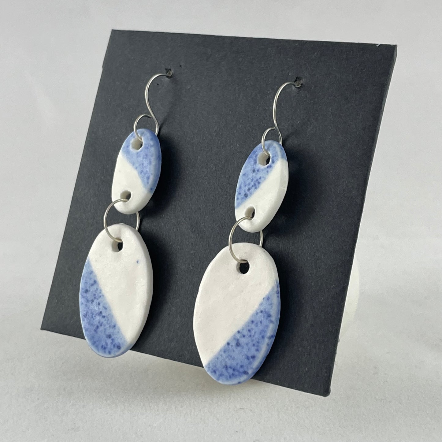 A pair of handmade porcelain earrings on a black background. Asymmetrical glaze pattern in blue and white on oval pendants. Side view