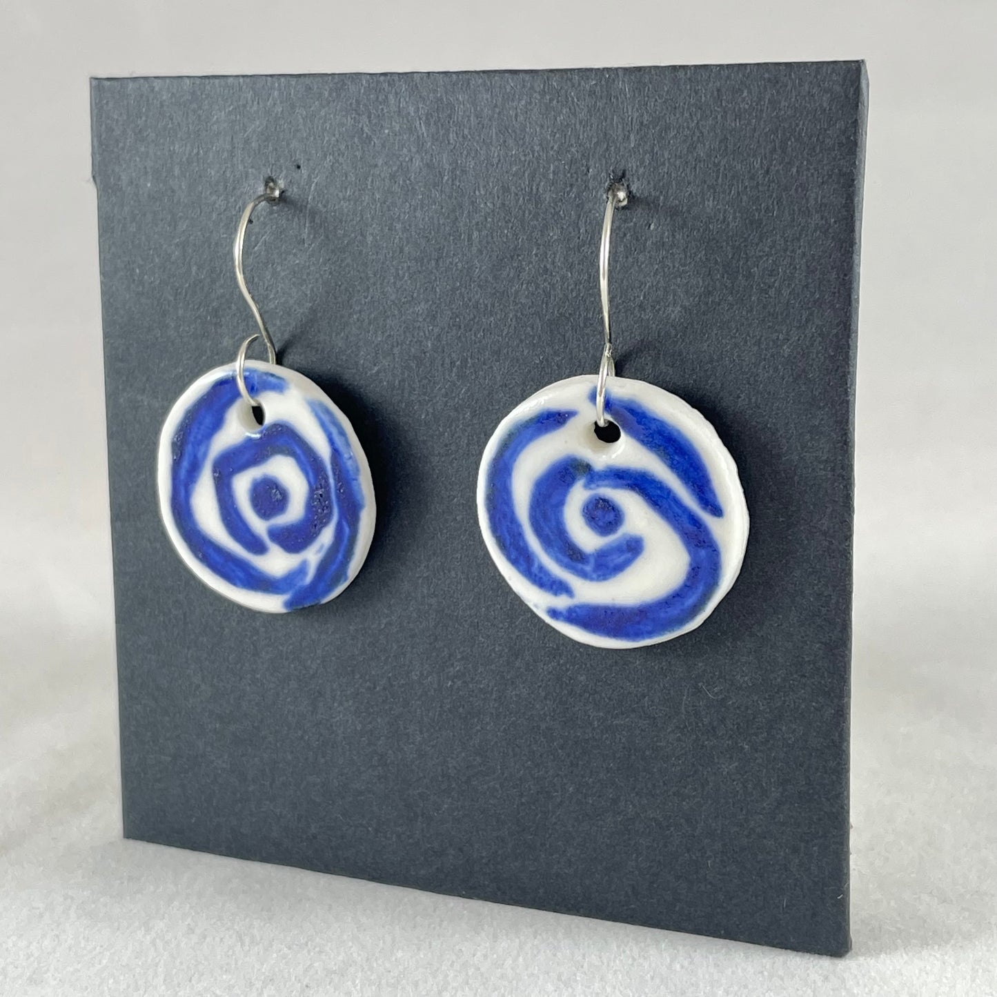 A pair of blue and white porcelain earrings hanging on black card. Loosely painted blue circles on white porcelain. Photographed from left hand side showing slim earring profile.