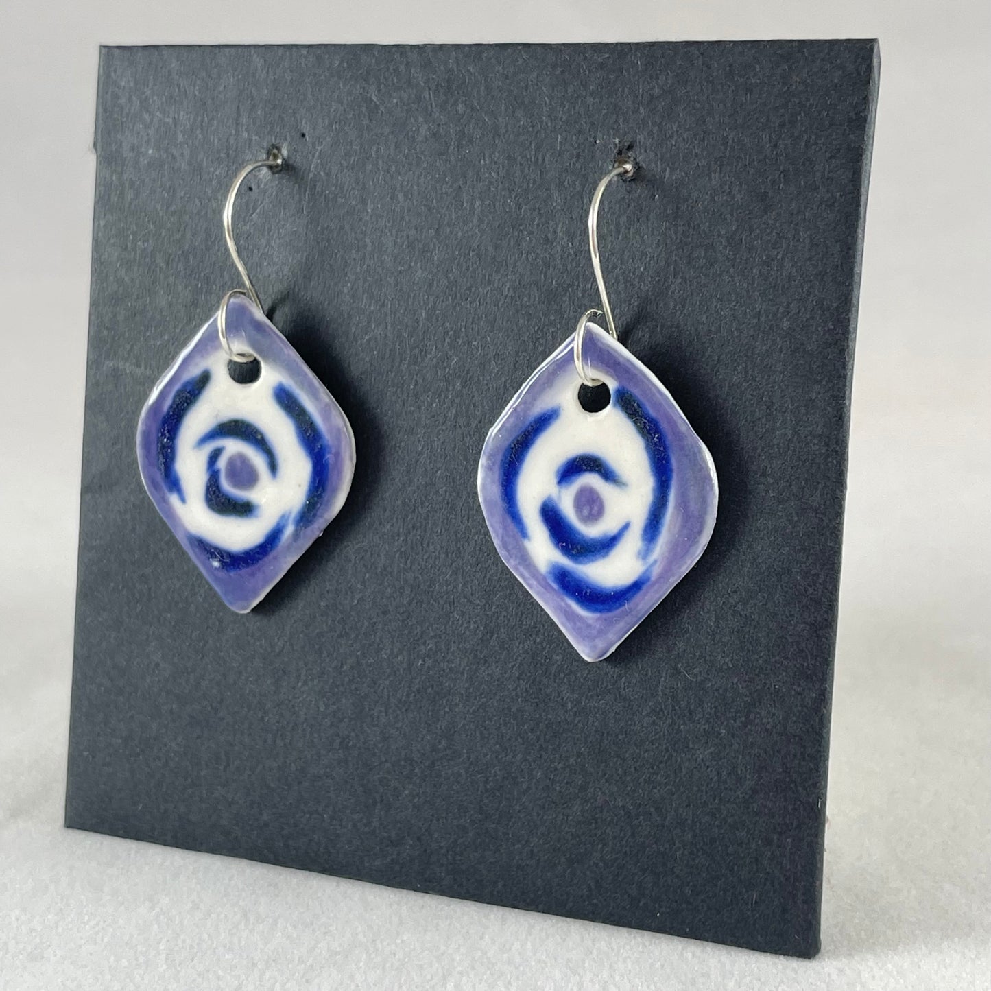 A pair of lavender, blue and white porcelain earrings hanging on black card. Loosely painted blue circles on white porcelain with lavender border. Photographed from left hand side showing slim earring profile