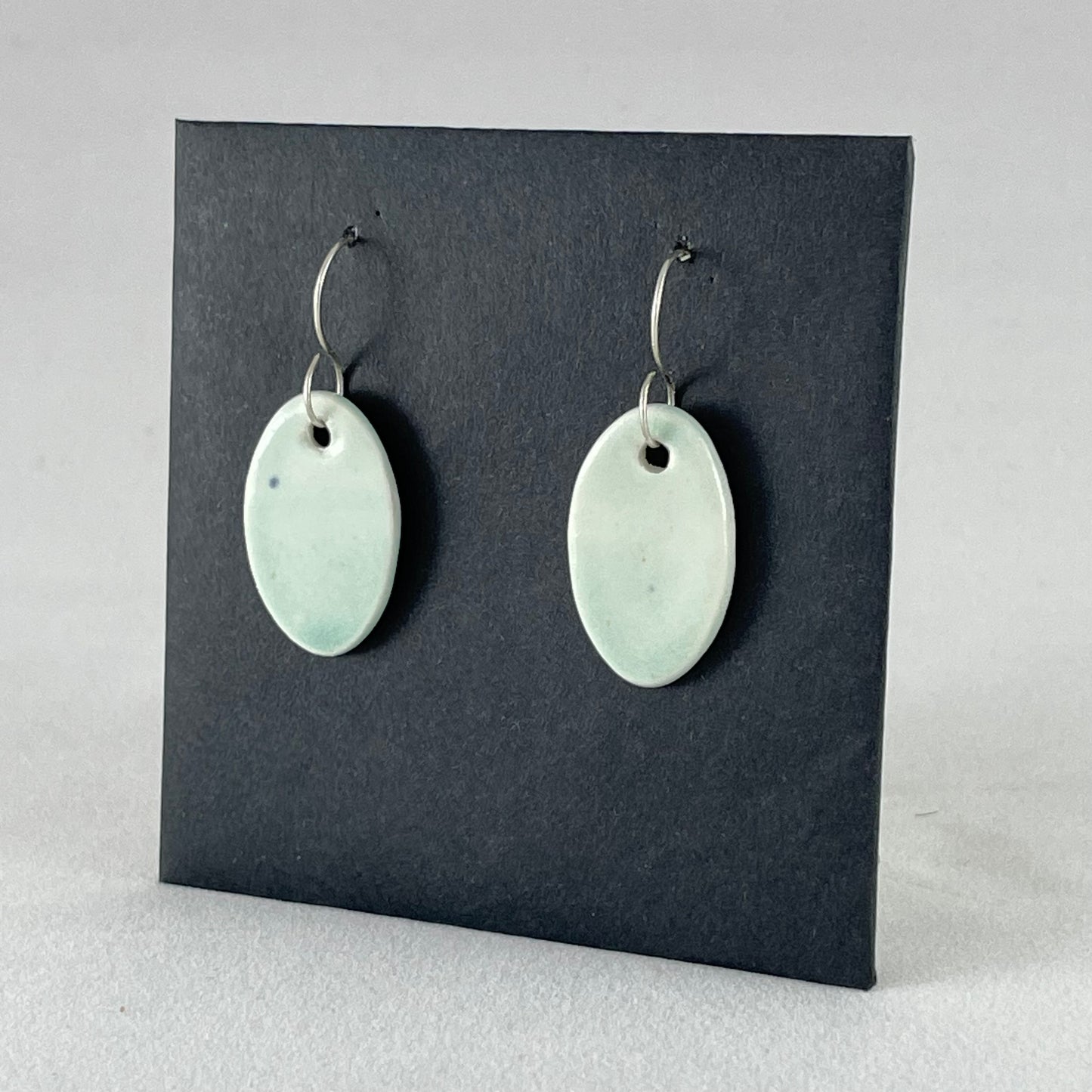 Pair of very pale mint green oval shaped porcelain earrings. Pictured in front of black background. Side view