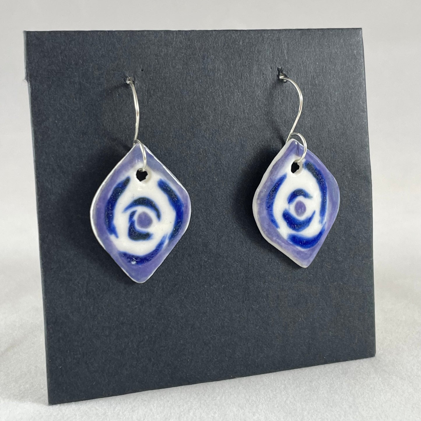 A pair of lavender, blue and white porcelain earrings hanging on black card. Loosely painted blue circles on white porcelain with lavender border. Photographed from right hand side showing slim earring profile