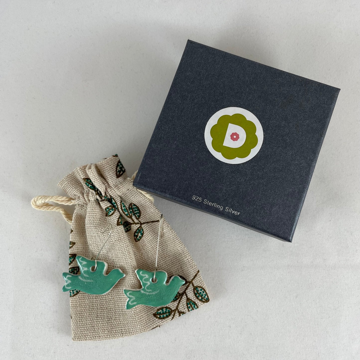A pair of green porcelain earrings in the shape of flying dove. Photographed on a linen pouch next to jewellery box.