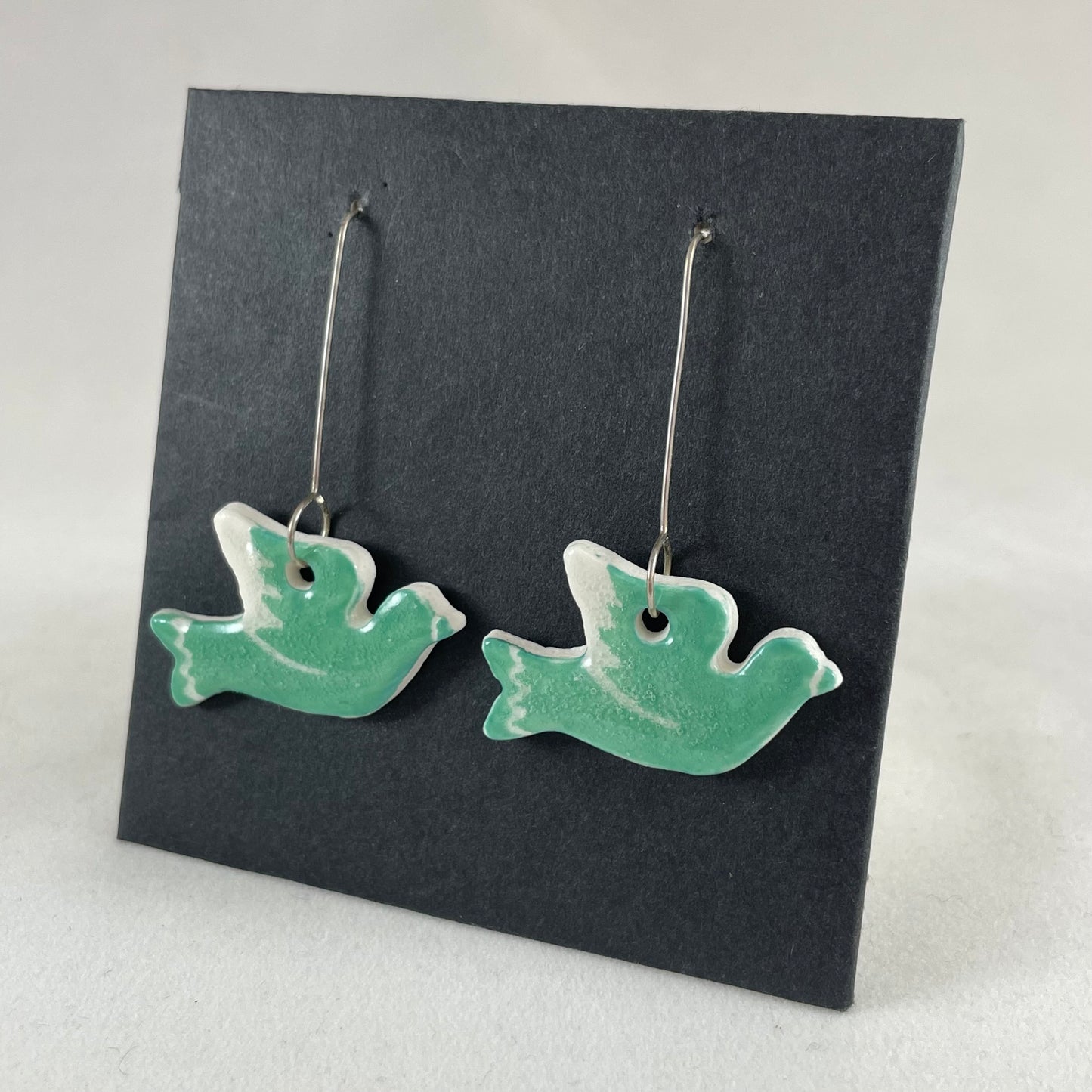 A pair of green porcelain earrings in the shape of flying dove on a black background. Side view