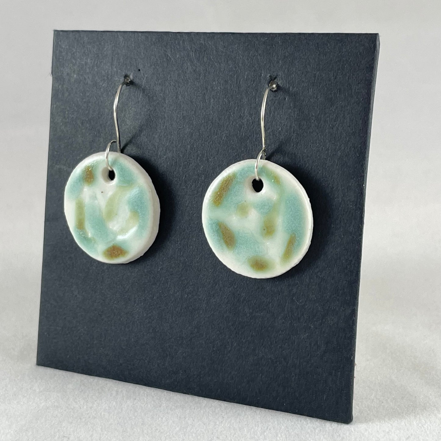 A pair of mint, olive green and white porcelain earrings hanging on black card. Photographed from right hand side showing slim earring profile.