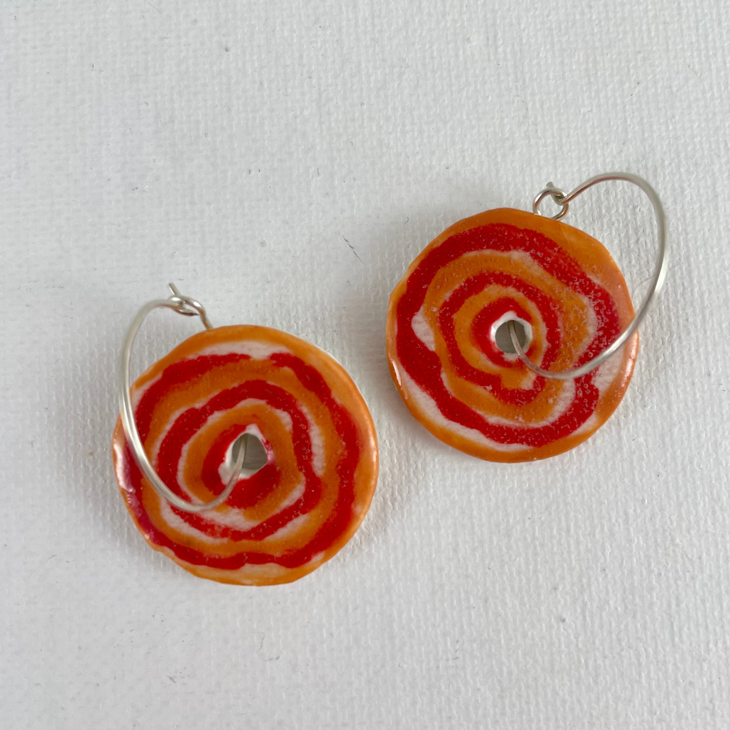 Closeup of pair of red and orange porcelain earrings on a white background. Simple rose pattern with alternating red and orange circles loosely painted. Small visible areas of white porcelain.