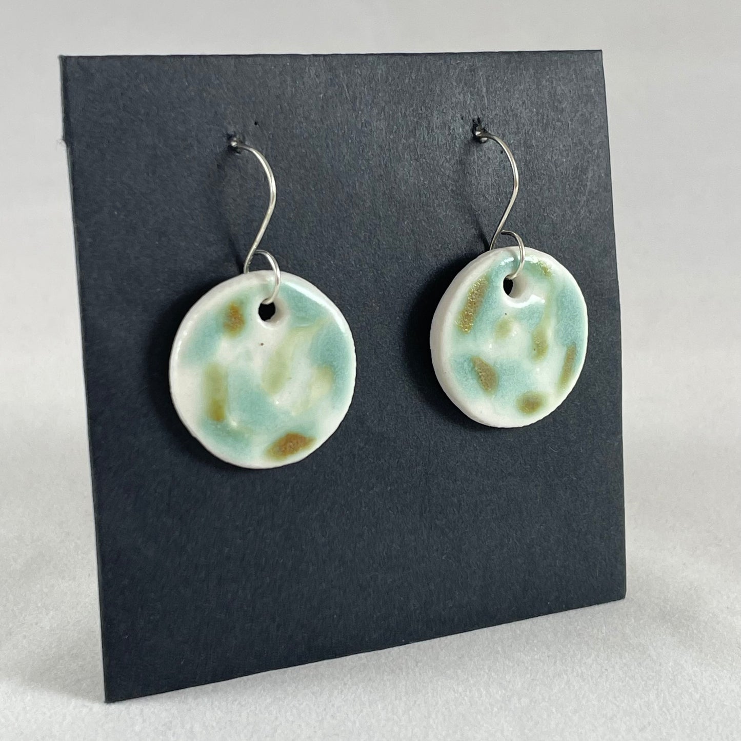A pair of mint, olive green and white porcelain earrings hanging on black card. Photographed from left hand side showing slim earring profile.