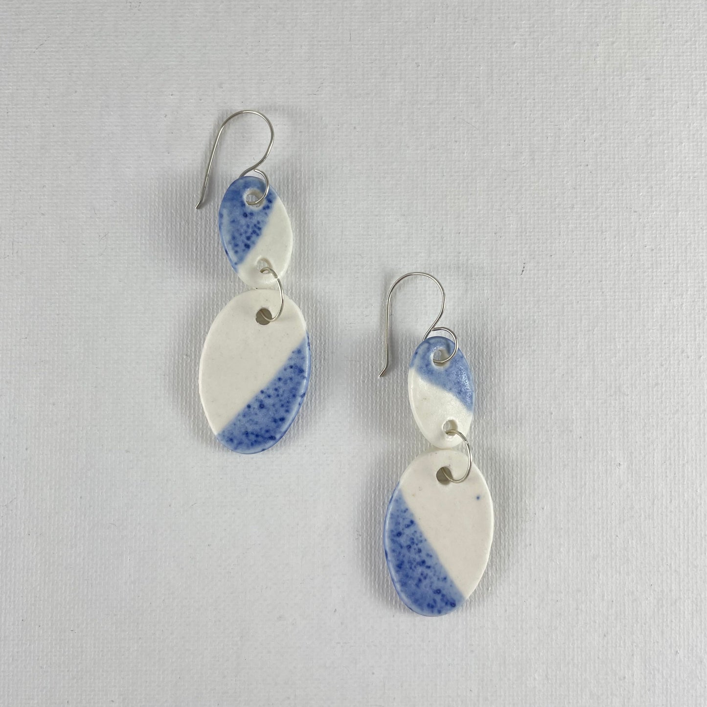 A pair of handmade porcelain earrings laying flat on a white background. Asymmetrical glaze pattern in blue and white on oval pendants.