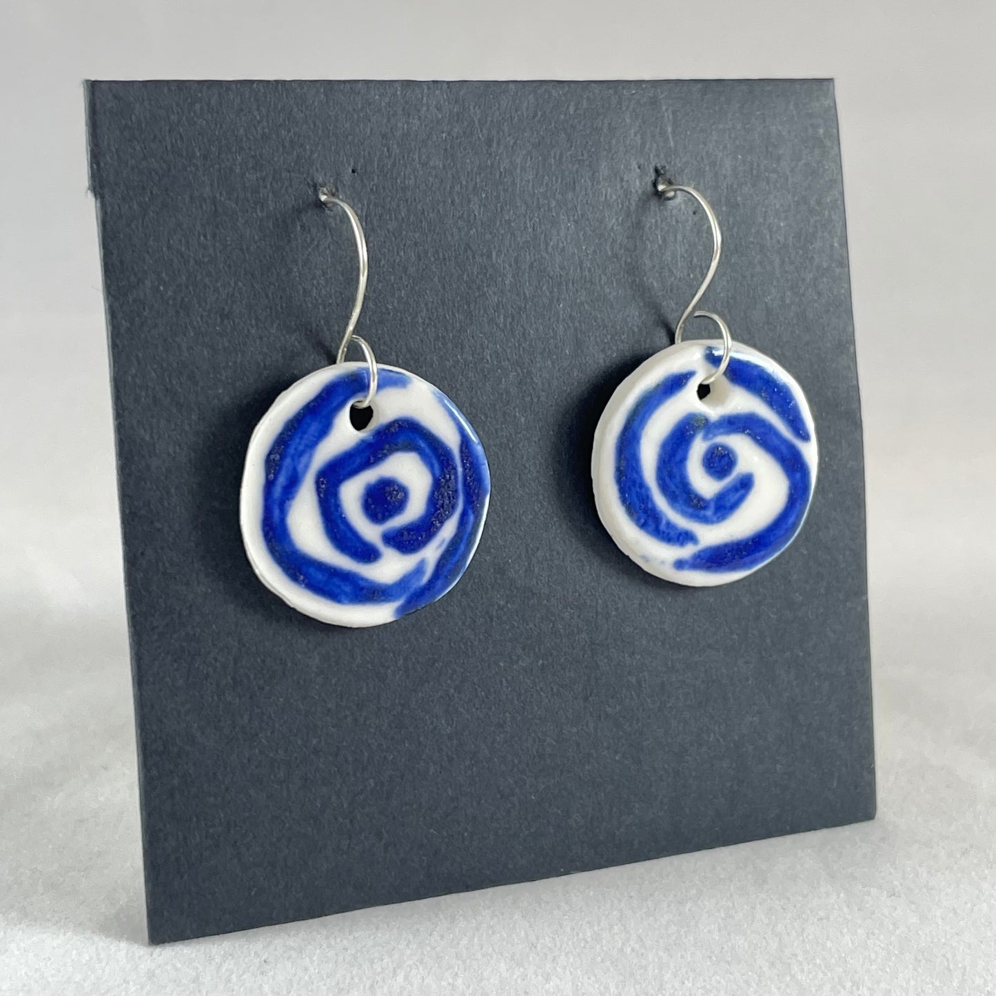 A pair of blue and white porcelain earrings hanging on black card. Loosely painted blue circles on white porcelain. Photographed from right hand side showing slim earring profile.