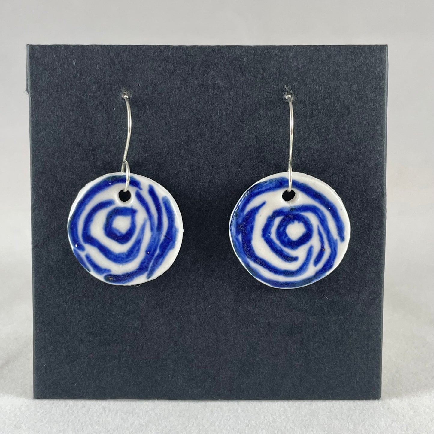 A pair of blue and white porcelain earrings hanging on black card. Simple rose pattern with loosely painted blue circles on white porcelain.