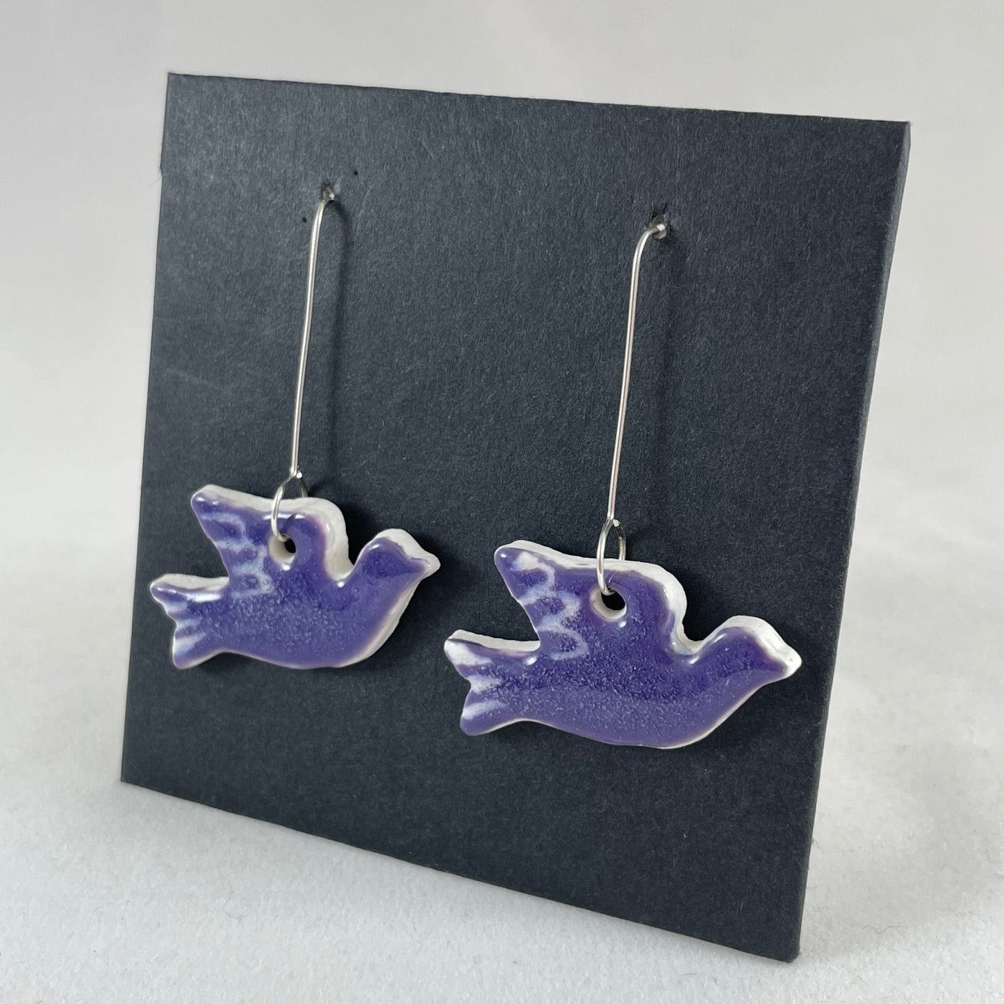 A pair of lavender porcelain earrings in the shape of flying dove on a black background. Side view