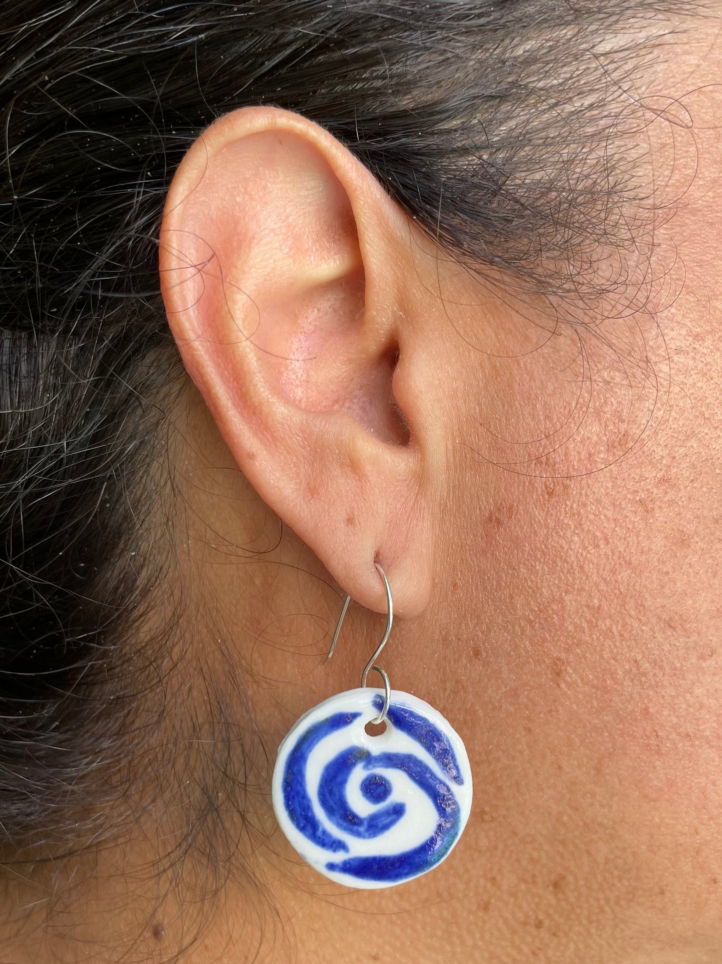 Closeup of woman with tan skin wearing white and blue circular earrings with rose pattern. Woman has her dark brown hair is pulled back in ponytail.