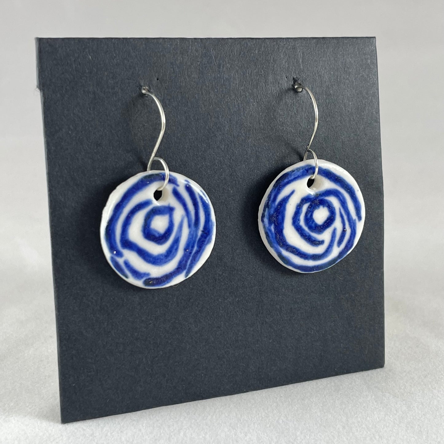 A pair of blue and white porcelain earrings hanging on black card. Simple rose pattern with loosely painted blue circles on white porcelain. Photographed to show earrings from left hand side