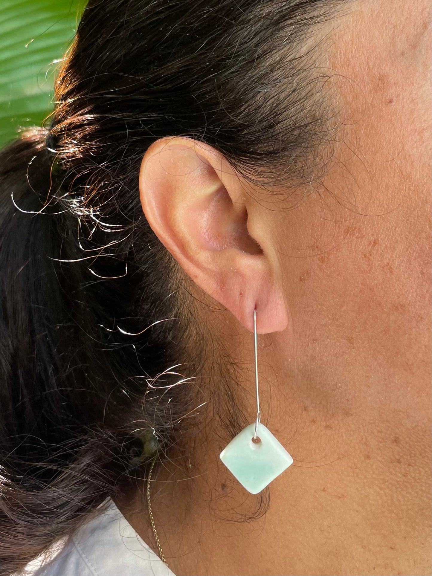 Closeup of woman wearing pale mint green earrings. Woman has olive skin and her dark brown hair is pulled back in ponytail. Sun is shining on her hair