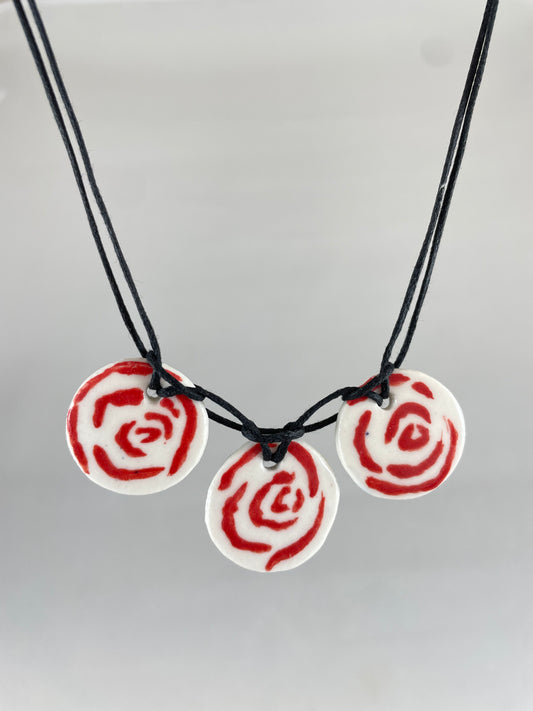 Necklace with three circular porcelain pendants on black coloured thread, hanging in front of a white background. Red rose pattern painted on white porcelain.