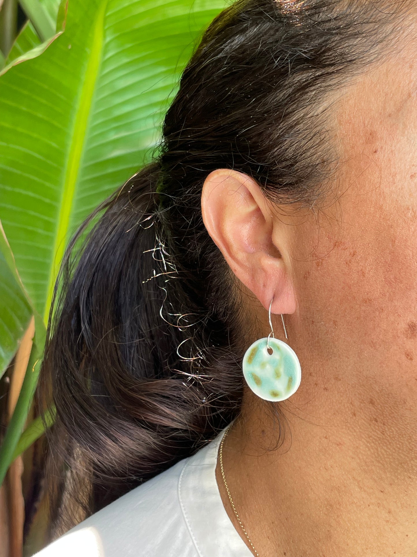Closeup of woman with tan skin wearing mint, olive green and white and circular earrings. Woman has her dark brown hair is pulled back in ponytail.