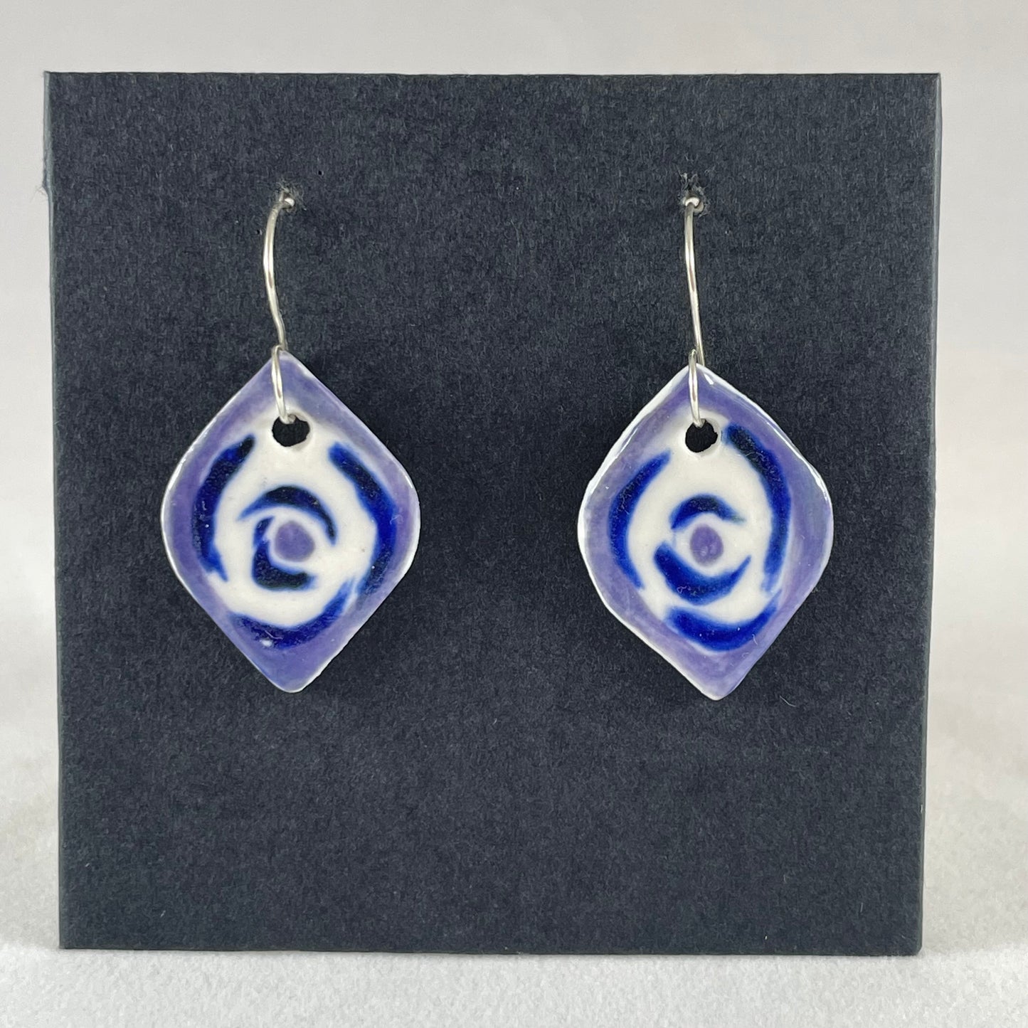 A pair of blue, lavender and white porcelain earrings hanging on black card. Pointed oval shape. Loosely hand painted blue circles on white porcelain, with a lavender border.