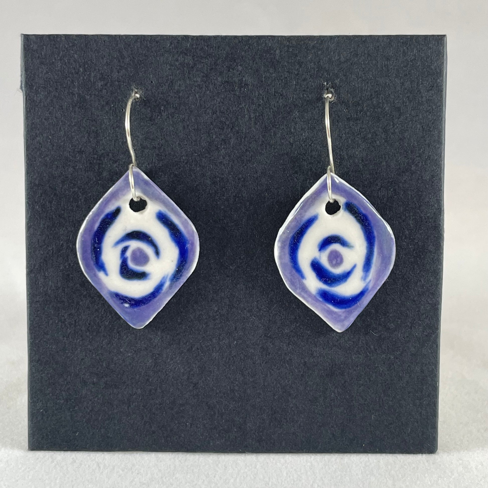 A pair of blue, lavender and white porcelain earrings hanging on black card. Pointed oval shape. Loosely hand painted blue circles on white porcelain, with a lavender border.