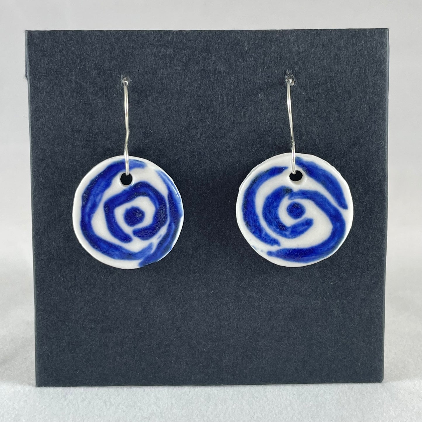A pair of blue and white porcelain earrings hanging on black card. Simple rose pattern with loosely painted blue circles on white porcelain.
