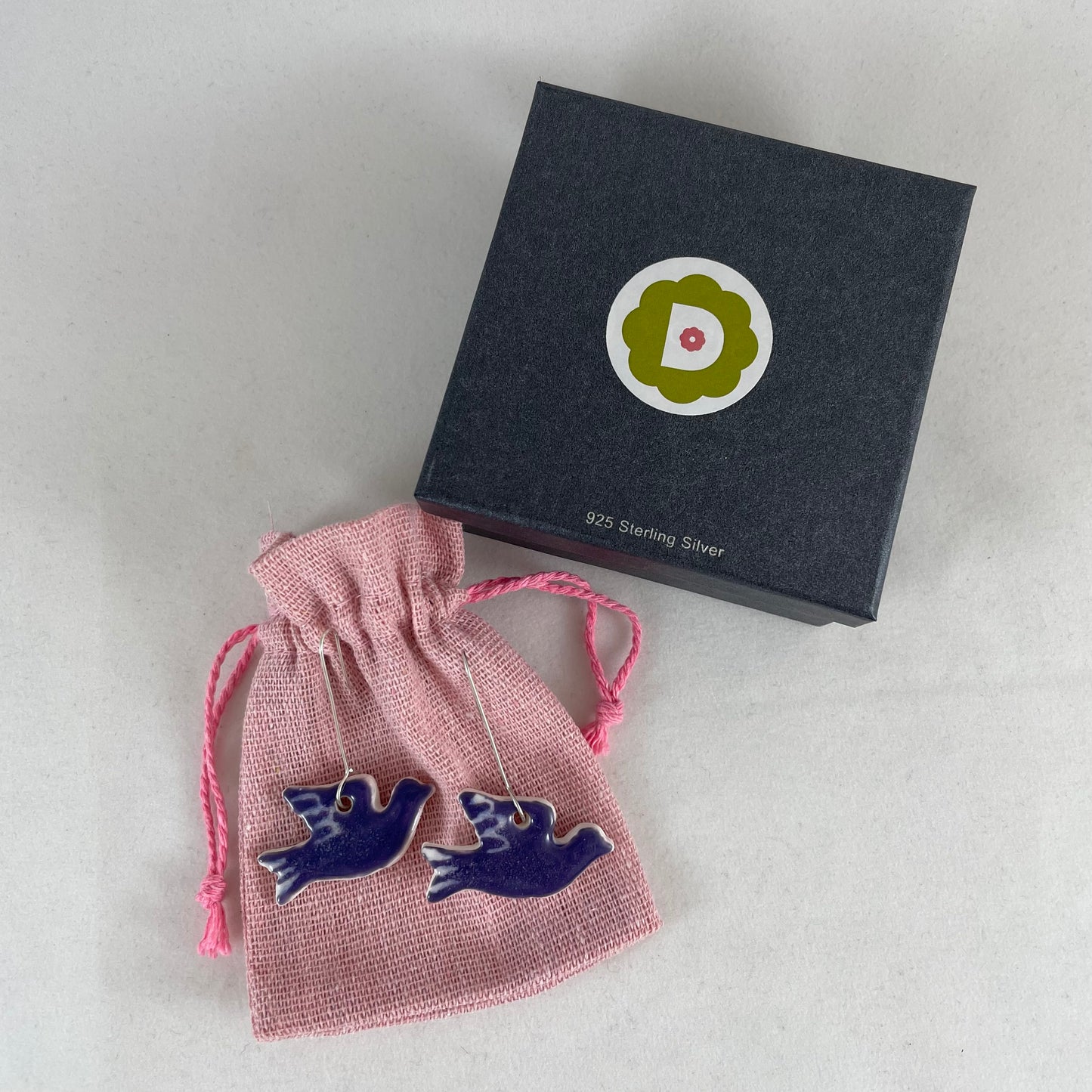A pair of lavender porcelain earrings in the shape of flying dove. Photographed on a linen pouch next to jewellery box.