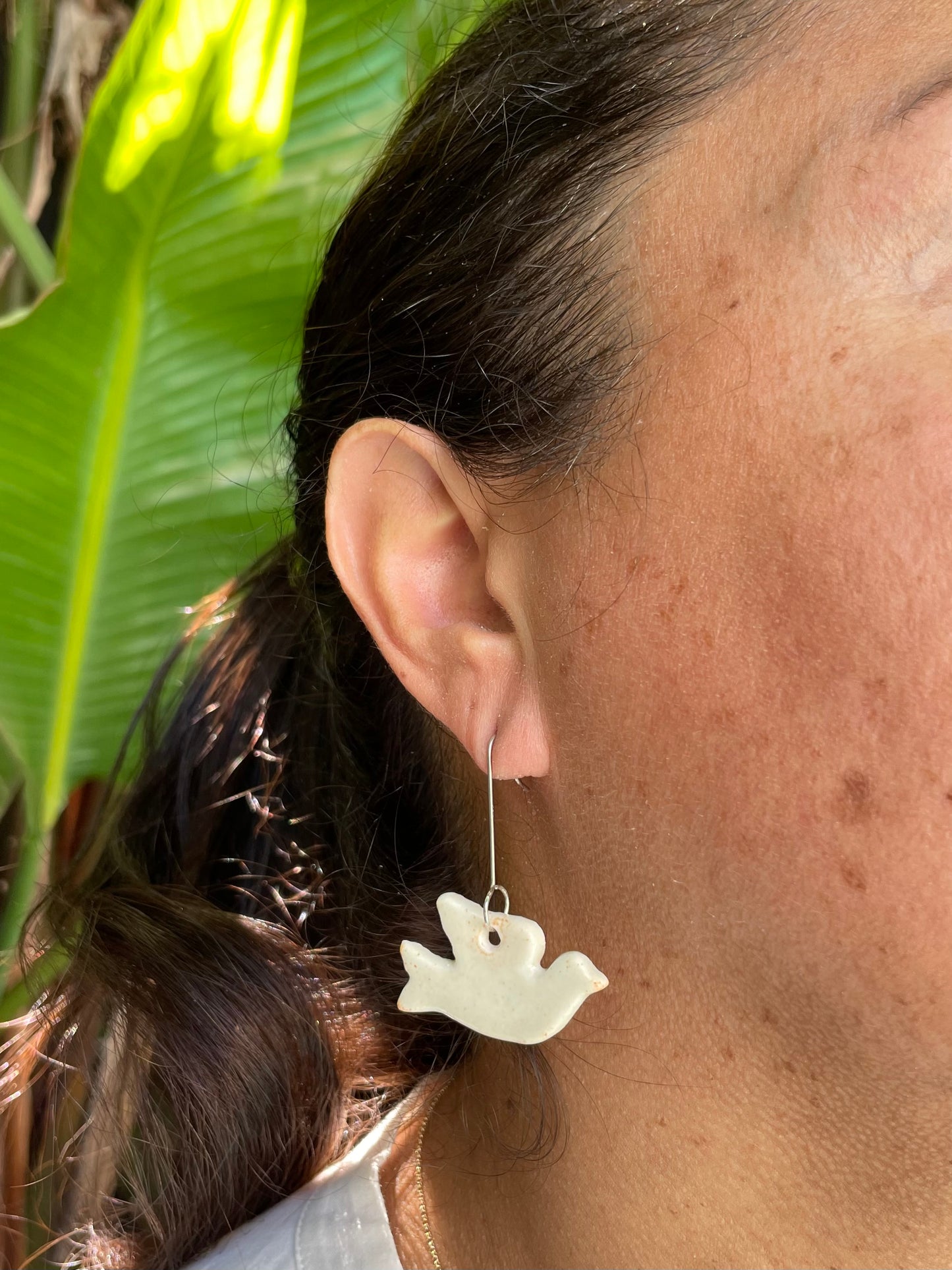 Closeup of woman wearing cream dove earrings. Woman has freckled olive skin and her dark brown hair is pulled back in ponytail. Sun is shining in garden