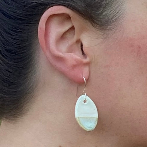Closeup of ear wearing an oval-shaped earring on fine silver hook. Asymmetrical pattern with half white glaze and half pale yellow and blue glaze.