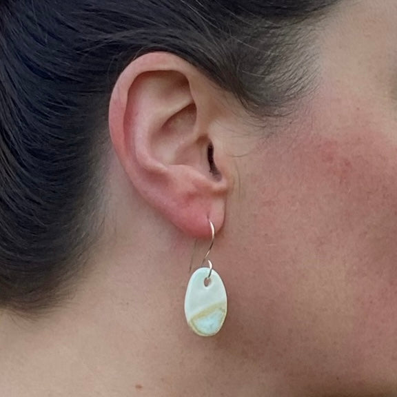 Close-up of an ear wearing an oval shaped earring on fine silver hook. Asymmetrical pattern with half white glaze and half pale yellow and blue glaze.